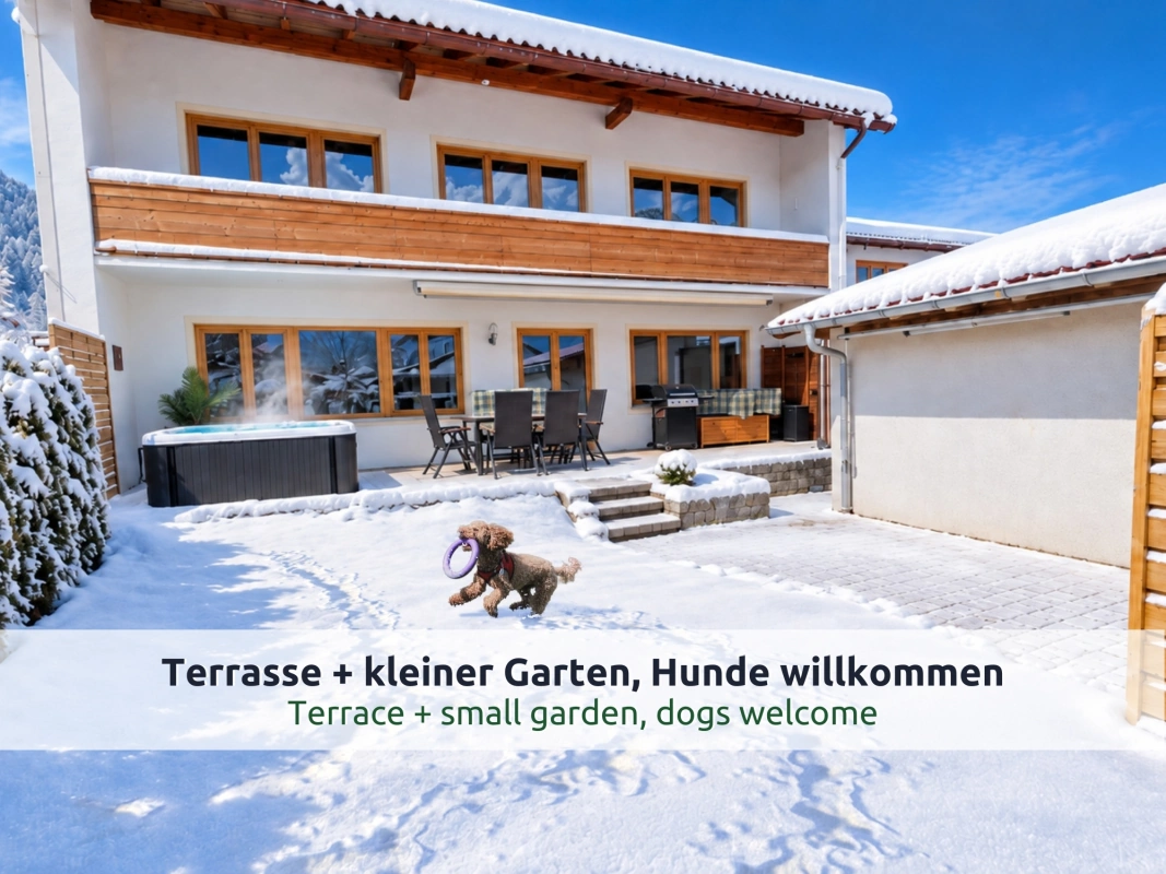 03 Ferienhaus Ruhpolding im Winter: Haus mit Holzbalkon, verschneiter Garten mit spielendem Hund und Terrasse mit Whirlpool