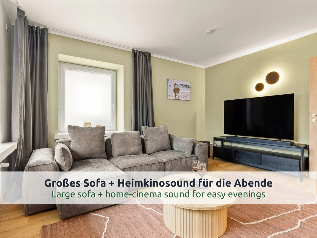 Ferienhaus Ruhpolding: Wohnzimmer mit grauem Ecksofa, großem Fernseher auf schwarzem Lowboard, rundem Holztisch, rosafarbenem Teppich und grün gestrichenen Wänden