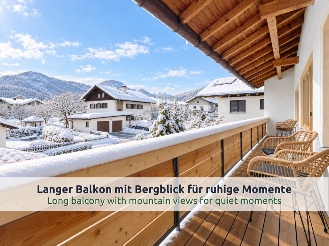 06 Ferienhaus Ruhpolding: langer Holzbalkon im Winter mit drei Rattanstühlen und Blick über verschneite Dächer und Gärten auf schneebedeckte Berge
