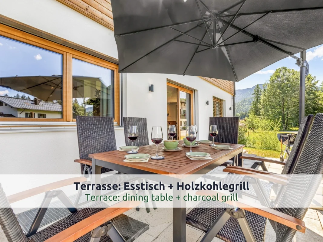 Chalet Reit im Winkl: Terrasse mit Holztisch, Geflechtstühlen und großem Sonnenschirm, gedeckter Tisch mit Weingläsern, Blick auf Wiese, Bäume und Berge