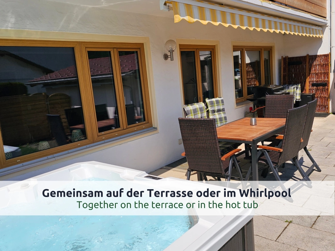 Ferienhaus Ruhpolding: Sommerterrasse mit privatem Whirlpool, Holztisch und Geflechtstühlen mit karierten Kissen, Palme und großen Holzfenstern zum Wohnzimmer