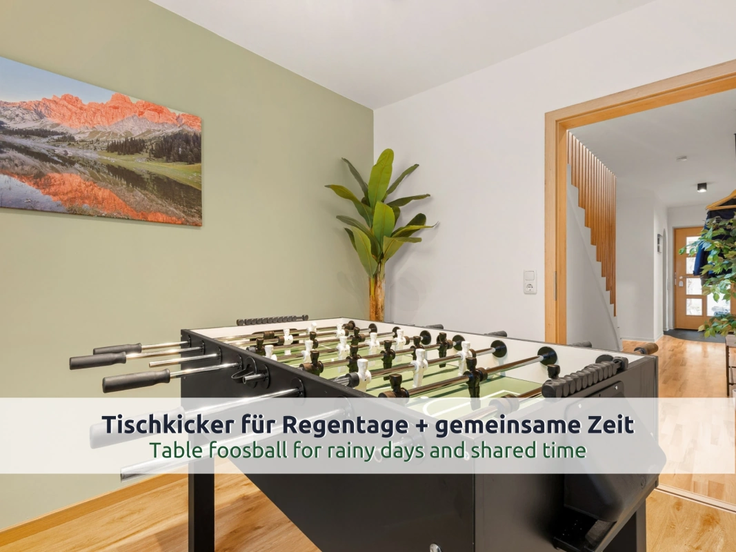 19 Ferienhaus Ruhpolding: Tischkicker im Spielzimmer mit grüner Wand, Bergpanorama-Bild und Zimmerpflanze; durch die Tür Blick in den Flur mit Holztreppe