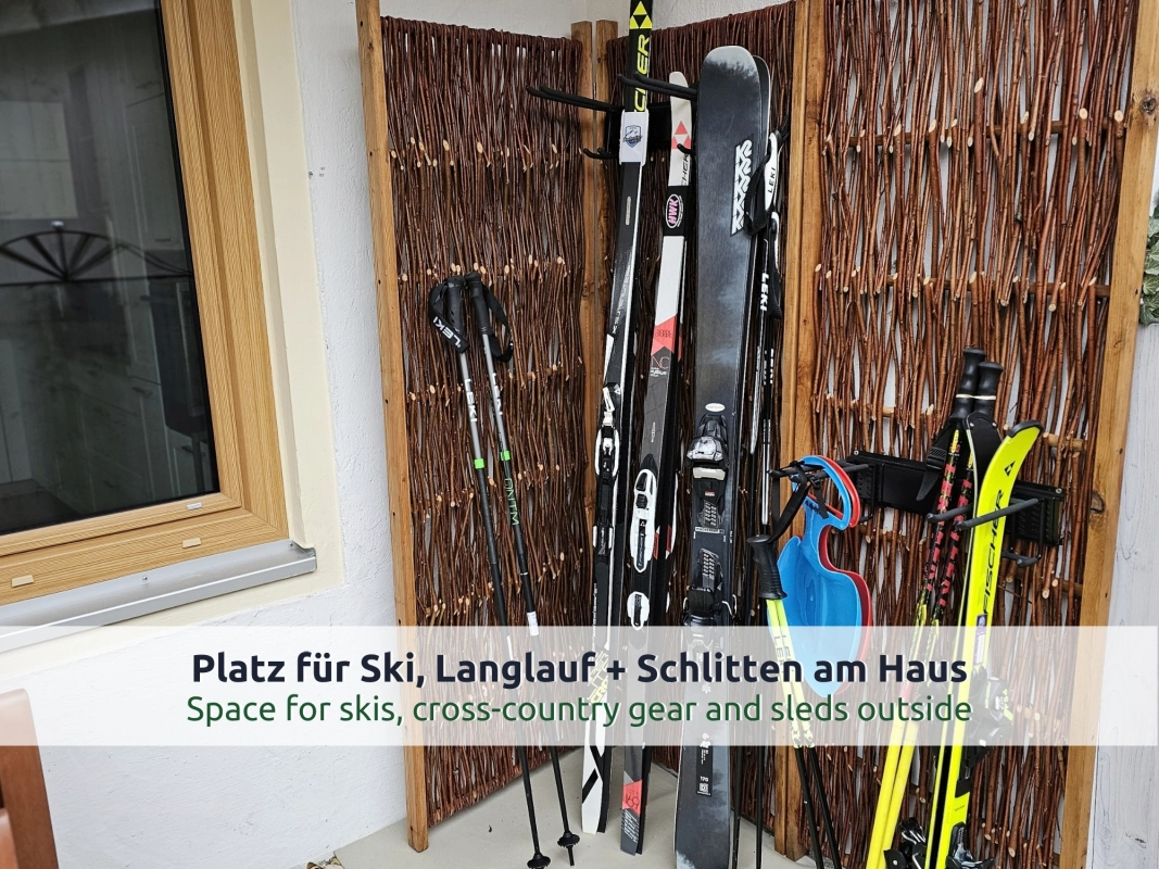 Ferienhaus Ruhpolding: Ski, Skistöcke und ein blauer Schneerutscher an einer Wand aus Weidengeflecht neben einem Fenster