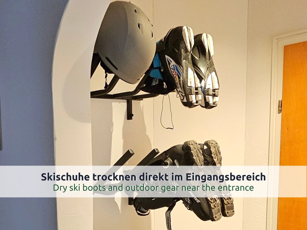 23 Ferienhaus Ruhpolding: Skihelm und zwei Paar Skischuhe an schwarzen Wandhaltern im Eingangsbereich, daneben eine orangefarbene Tür