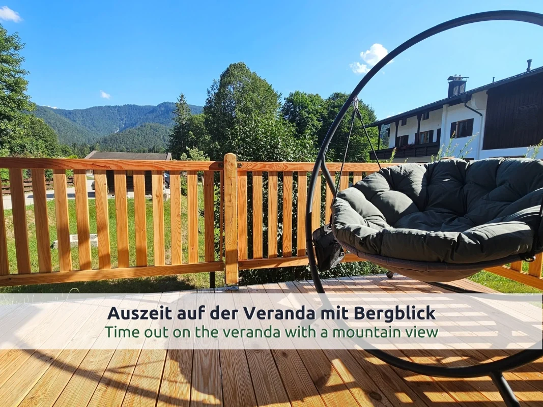 Chalet Reit im Winkl: Veranda mit rundem Hängesessel, Holzzaun, Wiese, Nachbarhaus und bewaldeten Bergen unter blauem Himmel