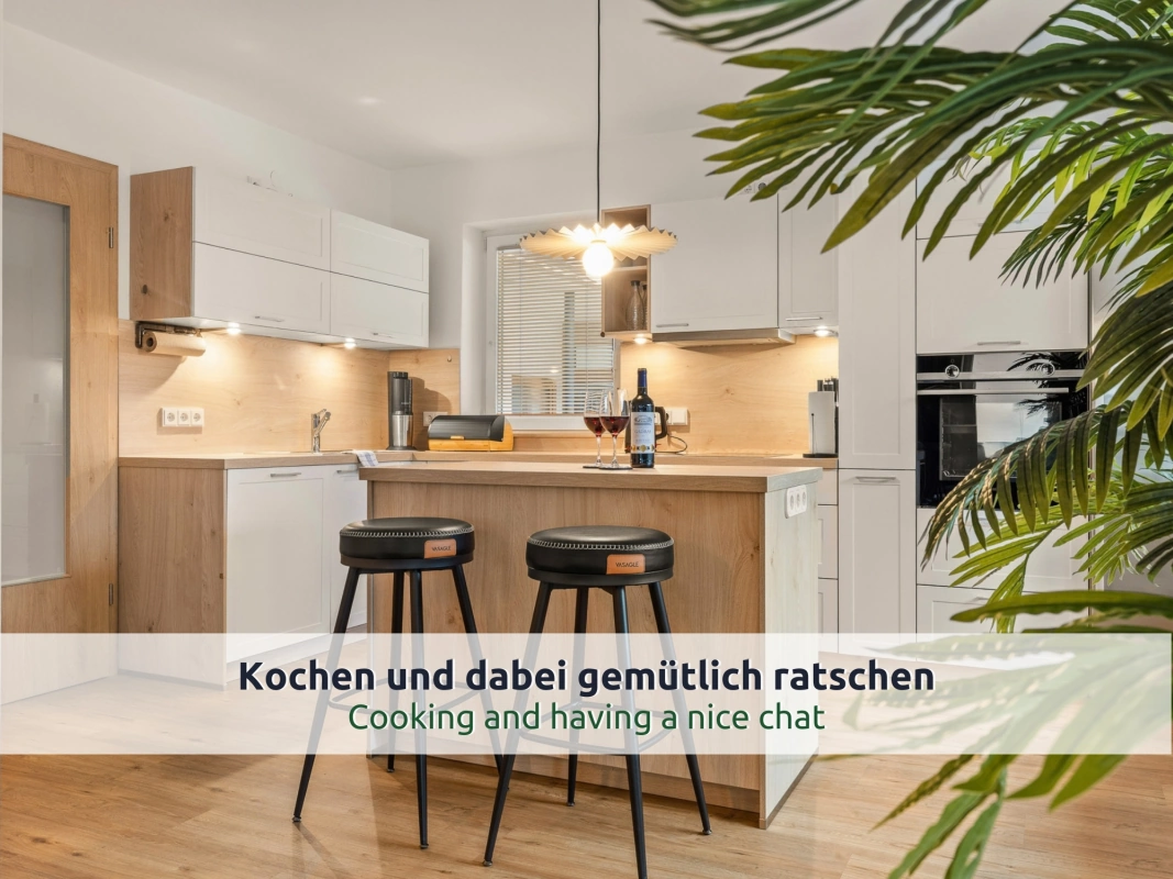 25 Ferienhaus Ruhpolding: Küche mit Kochinsel und zwei schwarzen Barhockern, weiße Hängeschränke, Holzrückwand, Pendelleuchte und eine Palme im Vordergrund