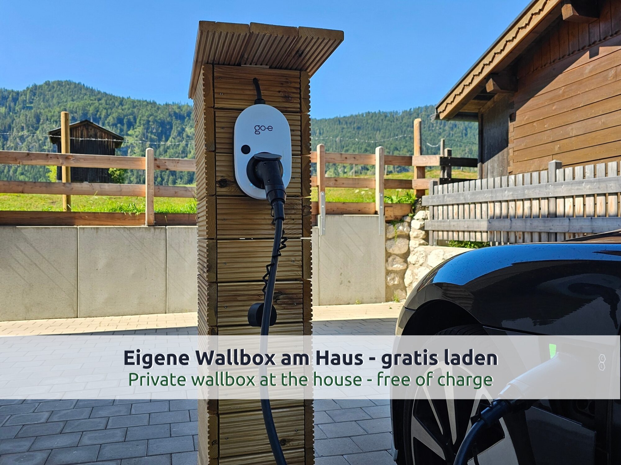 Chalet Reit im Winkl: Wallbox an einer Holzsäule mit Ladekabel an einem schwarzen Elektroauto, gepflasterte Einfahrt, Holzzaun und bewaldete Berge