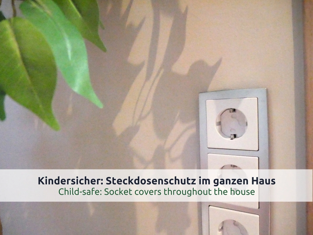 27 Ferienhaus Ruhpolding: Dreifachsteckdose mit eingesetzten Kindersicherungen an einer hellen Wand, Blätterschatten einer Zimmerpflanze