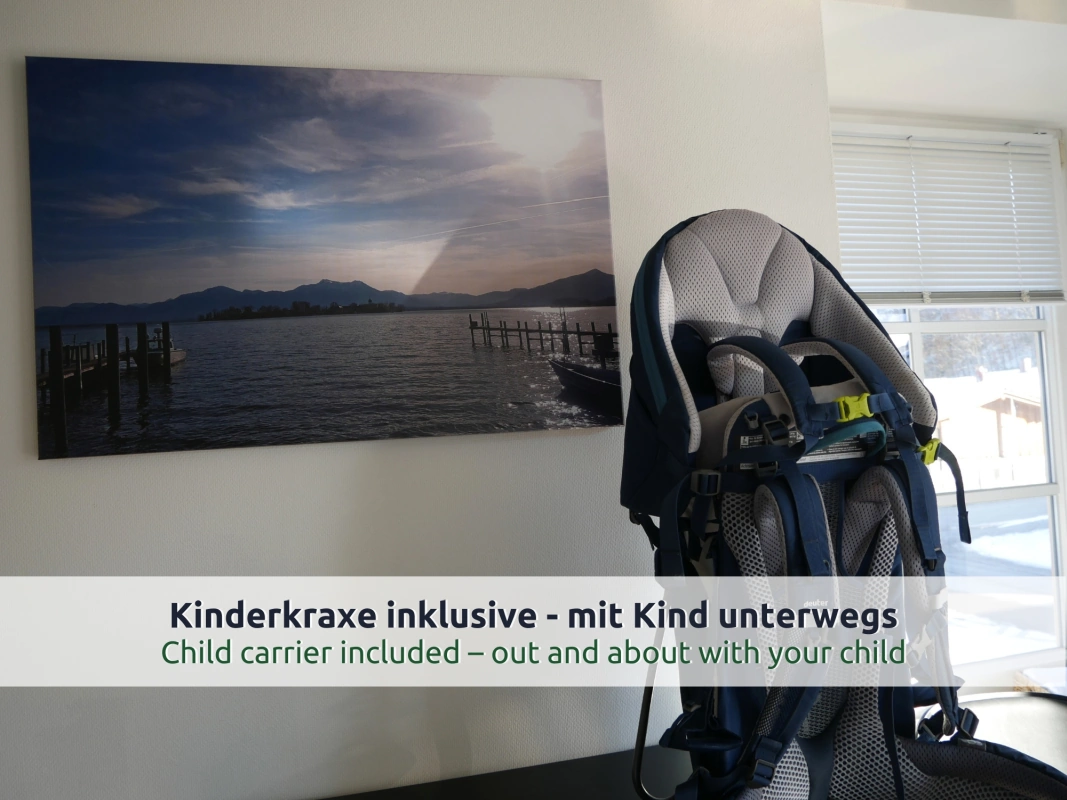 Ferienhaus Ruhpolding: Kindertrage-Rucksack in Grau und Blau auf einem Tisch, dahinter ein Seebild an der Wand und ein Fenster