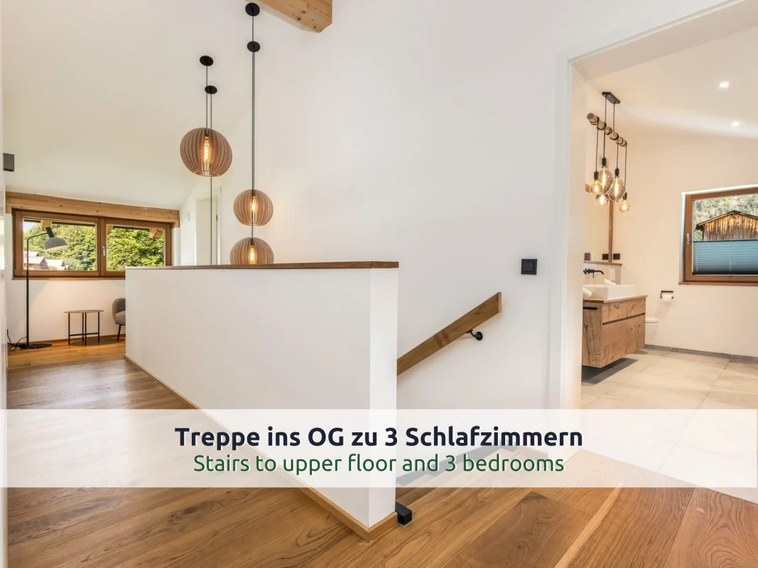 Chalet Reit im Winkl: Flur im Obergeschoss mit Treppenabgang, Holzhandlauf, Kugel-Pendelleuchten, Sitzecke am Fenster und offener Tür ins Bad