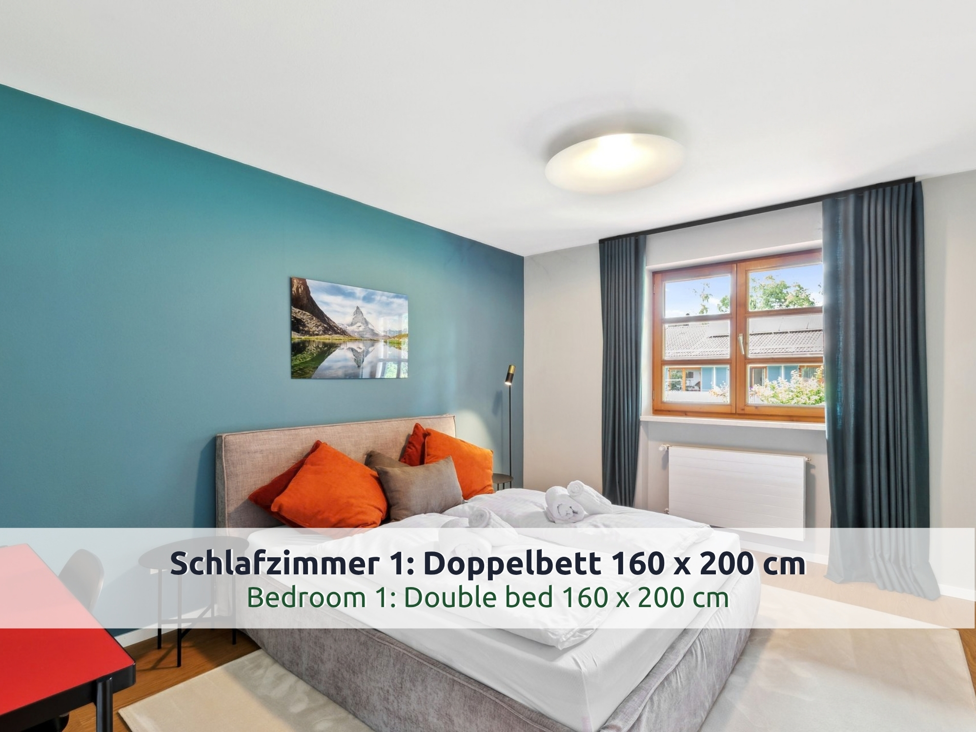 31 Villa Siegsdorf: Schlafzimmer mit Doppelbett, orangen Kissen, petrolfarbener Wand, Bergbild, Fenster mit dunklen Vorhängen und rotem Tisch am Rand