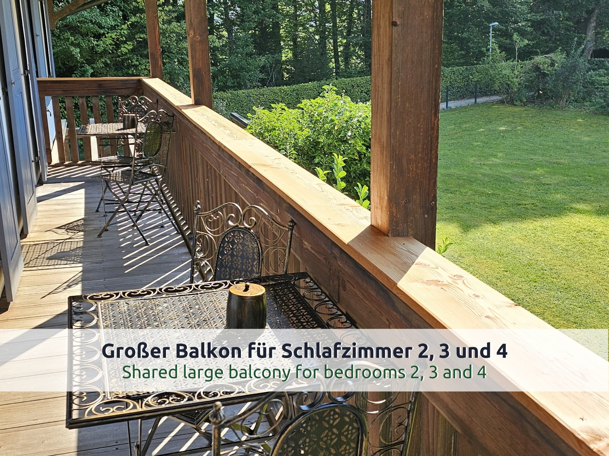 40 Villa Siegsdorf: Holzbalkon im Obergeschoss mit verschnörkeltem Metalltisch und Stühlen, Blick auf Rasen, Hecke und Bäume in der Sonne