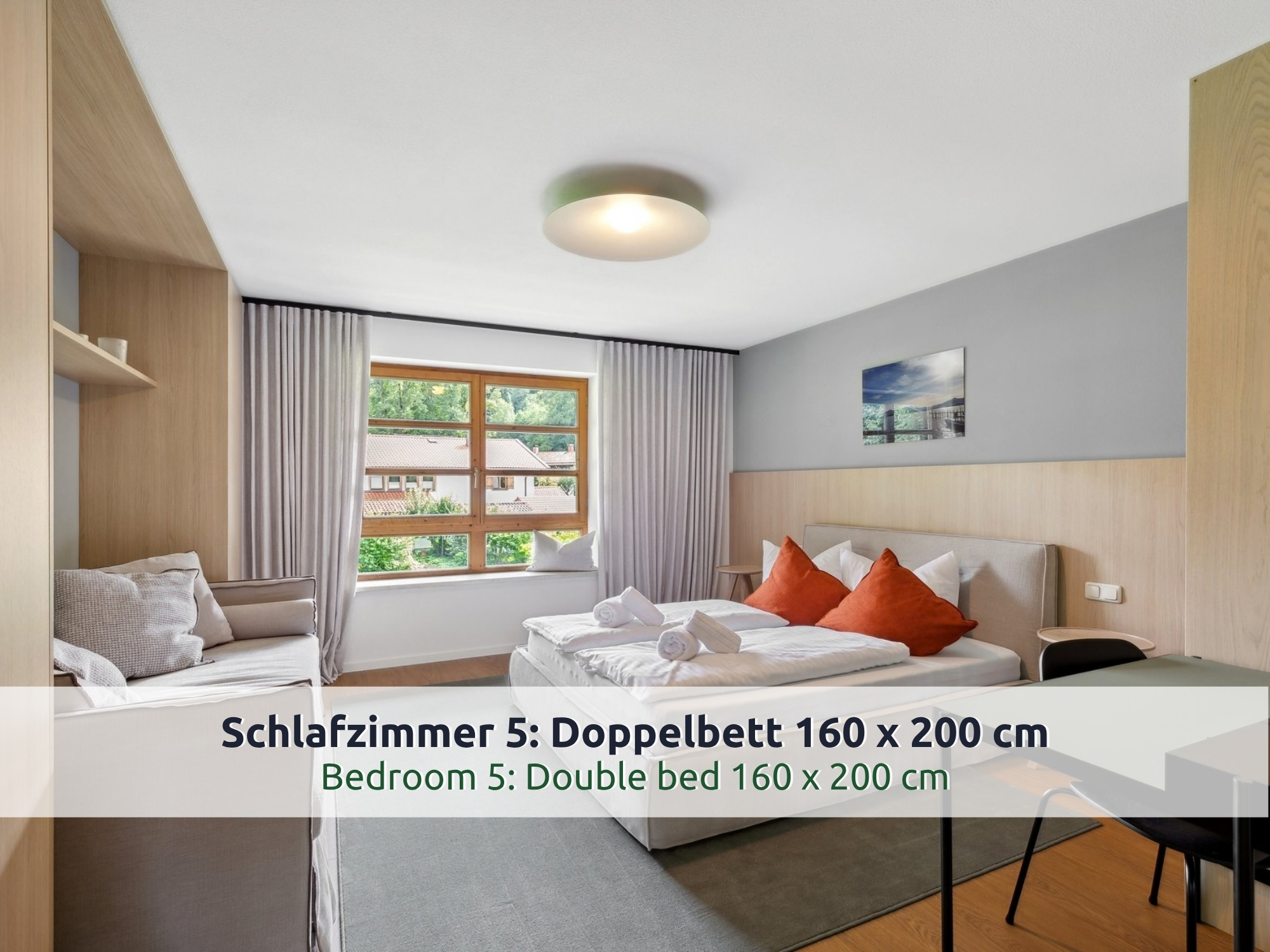 41 Villa Siegsdorf: großes Schlafzimmer mit Doppelbett, orangen Kissen, Holzverkleidung, hellem Sofa, Tisch mit Stuhl und Fenster mit Blick auf Dächer und Bäume