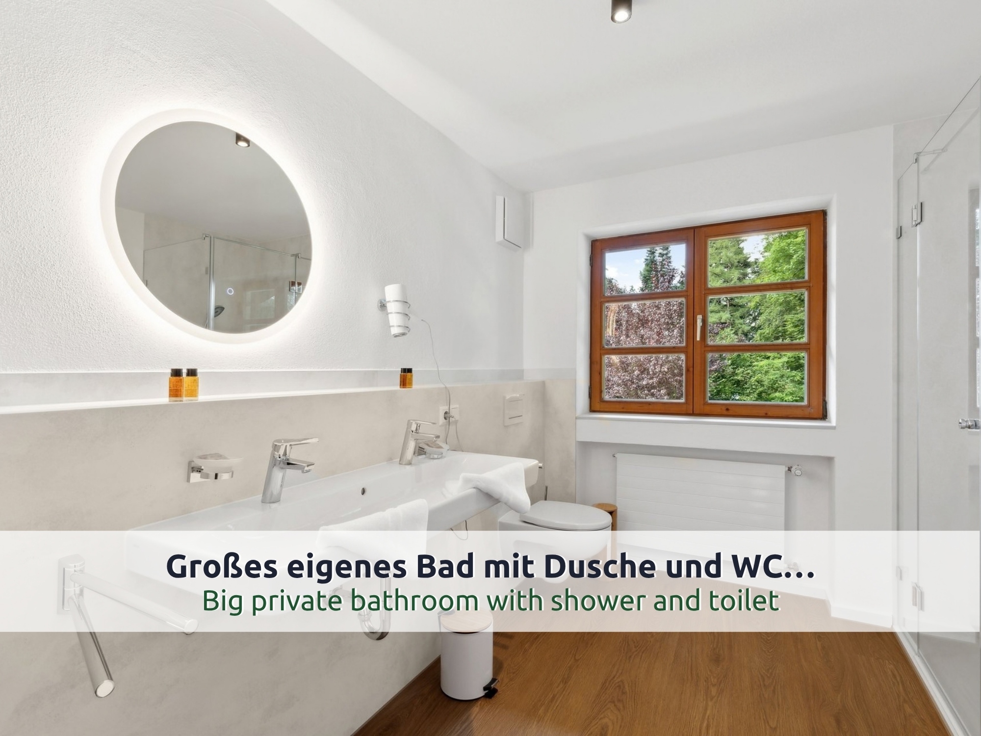 43 Villa Siegsdorf: helles Badezimmer mit Doppelwaschbecken, rundem beleuchtetem Spiegel, WC, Holzfenster und Glasdusche am rechten Rand