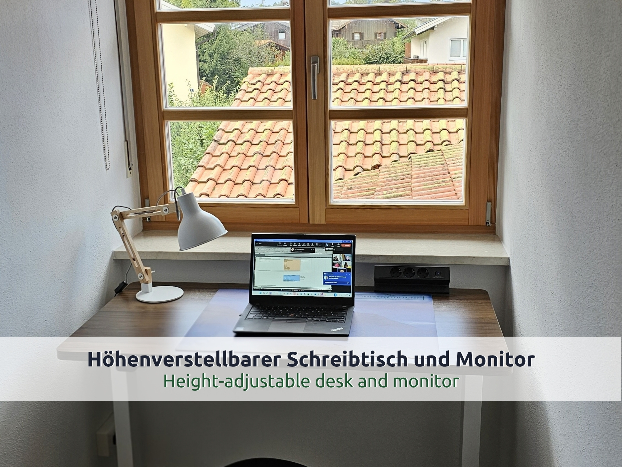 49 Villa Siegsdorf: Schreibtisch vor einem Holzfenster mit Blick auf Ziegeldächer, Laptop mit Videokonferenz, Schreibtischlampe und Steckdosenleiste