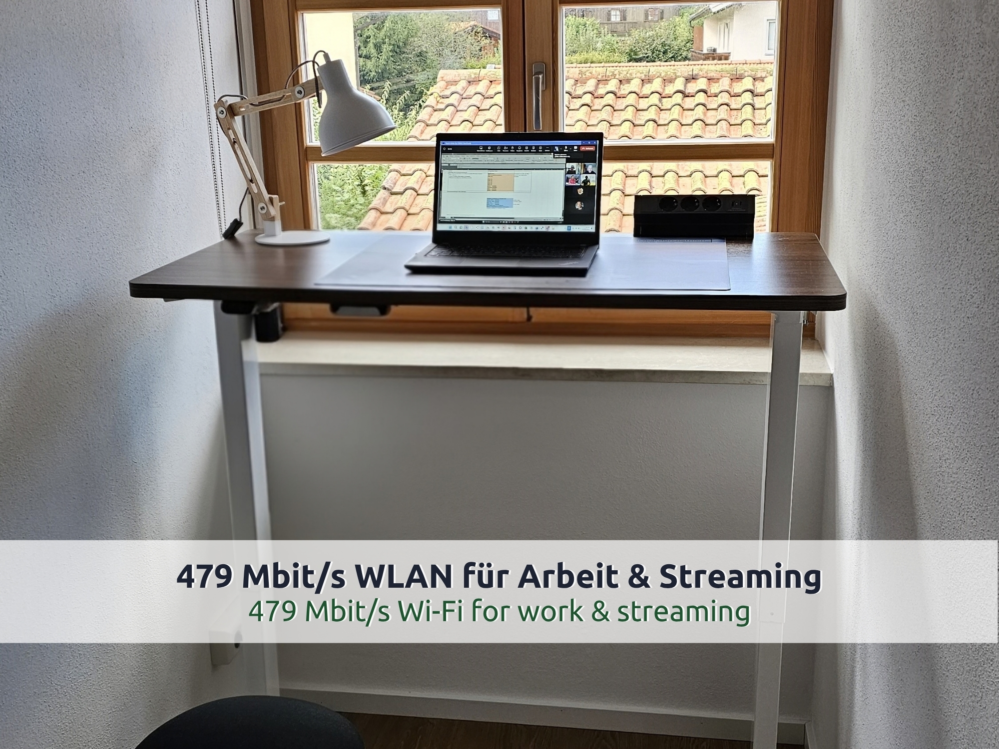 50 Villa Siegsdorf: höhenverstellbarer Schreibtisch auf Stehhöhe vor einem Holzfenster, Laptop, Lampe und Steckdosenleiste, Blick auf Dächer